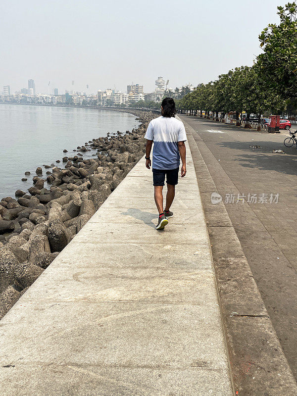 印度孟买，一名男子在海洋大道海堤上行走，混凝土防御，海岸管理，防止侵蚀，海水，c形长廊，装饰艺术建筑和摩天大楼排列在Girgaon Chowpatty公共海滩的后视图